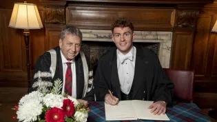 Image of Archie Goodburn sitting with Peter Mathieson signing principals medal document