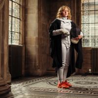 Georgia Haynes wearing graduate gown and fencing kit holding sabre in McEwan Hall by stained glass windows