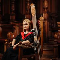 Emma Olley wearing graduate gown and sports kit sitting on red leather chair in McEwan Hall with skis propped against chair