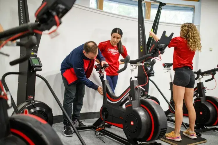 Two gym members adjusting an exercise bike whilst another works out