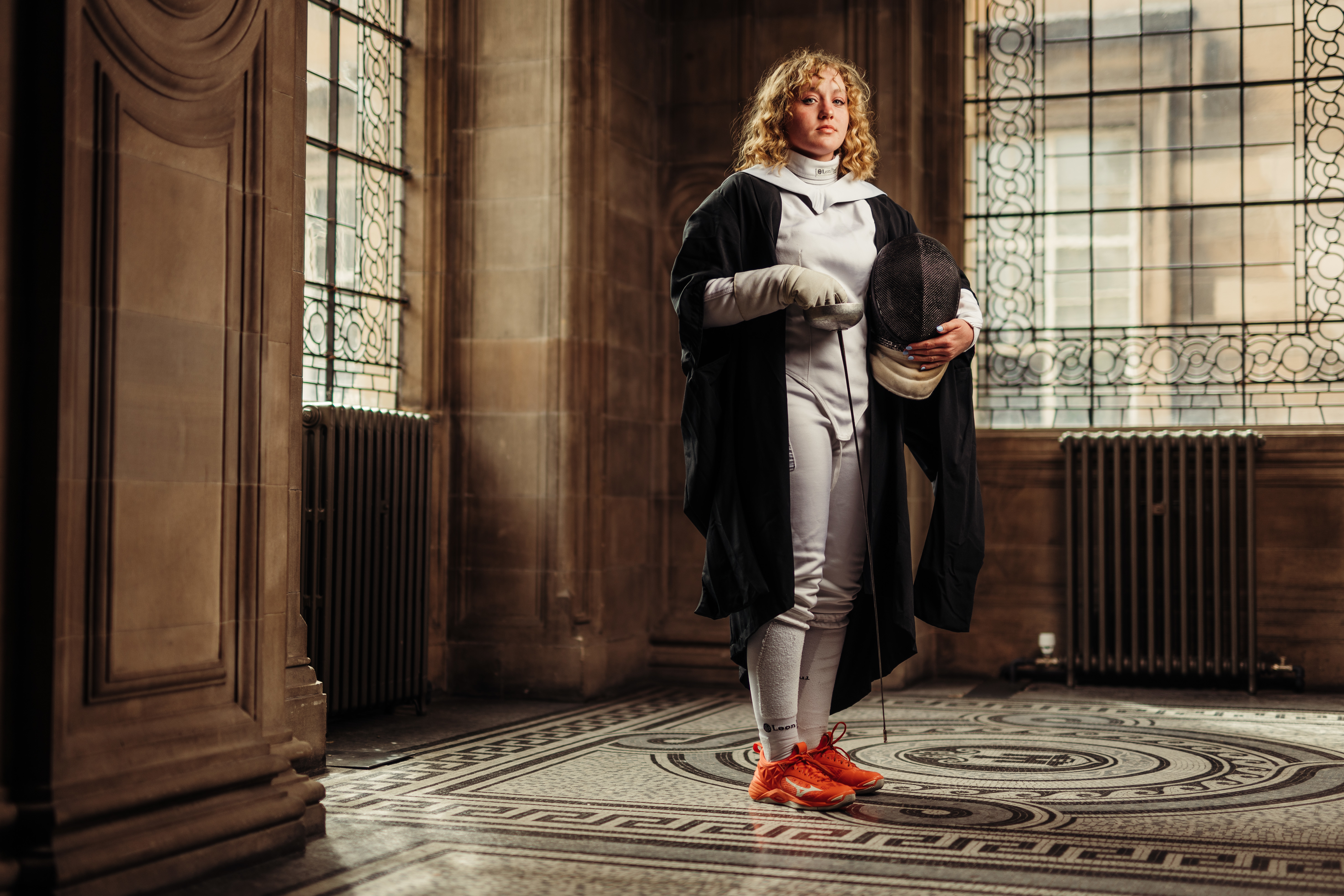 Georgia Haynes wearing graduate gown and fencing kit holding sabre in McEwan Hall by stained glass windows