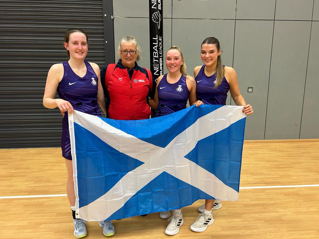 Image of netball players Holly Turnbull, Mia Mathewson and Katie Joslin with Head of Netball, Trish Wilcox holding scottish flag